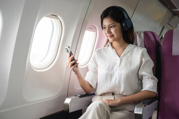 Asian woman using smartphone and headphones on airplane. Concept of air travel, technology, and in-flight entertainment
