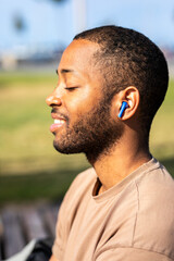 Fototapeta premium A joyful blind African young man basks in the sun at a park near the beach on a bright sunny day. He listens to music with headphones and happily responds to messages on his mobile phone.
