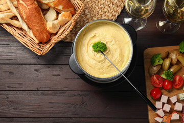 Fork with piece of broccoli, melted cheese in fondue pot, wine and snacks on wooden table, flat...