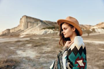 Woman in hat and blanket stands in deserted wilderness with majestic mountains in the distance