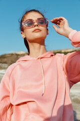 Stylish woman in pink sweatshirt and sunglasses standing confidently on sandy beach