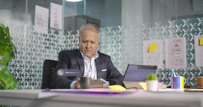 Indian Asian old aged adult gen x boss man sitting use tablet check bank loan crisis office place stress sad older elder fail male do over time work job feel bad upset talk phone call pop up notify