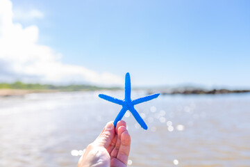 Hand holding a blue resin starfish. In the background, the sea shimmers with sunlight on the water. The image reflects the  and tropical climate