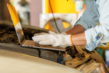 Mechanic's gloved hands working on a laptop placed on the car engine in a garage. Scene emphasizes...
