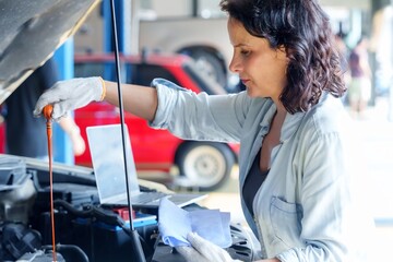 Female mechanic in blue denim shirt checking engine oil dipstick in a car engine, with laptop nearby in a garage. Demonstrates use of technology and manual inspection for automotive maintenance.