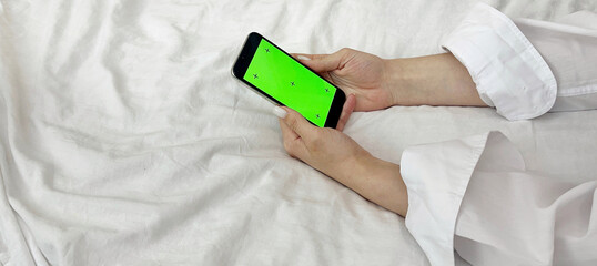 Female hands in a white shirt against the background of a bed hold a phone with a green screen. Mobile Phone Day. Mobile Phone Give Up Day