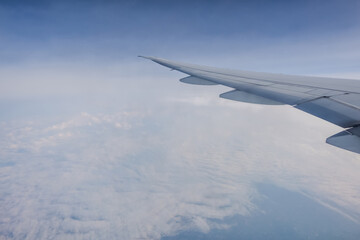View from an airplane window, showcasing the wing against a backdrop of a vast, cloud-filled sky. Scene conveys sense of height and tranquility, with the horizon blending into soft blue gradient.