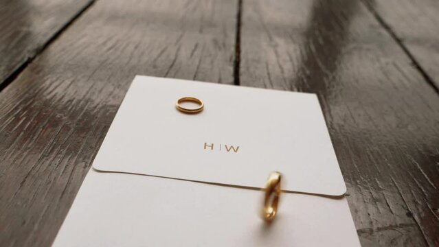 gold wedding rings spinning on invitation with initials H and W written in gold on wooden boards. Jewelry for a married couple. 60fps