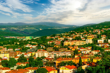 Fototapeta premium panoramic landscape view of green summer city with yellow houses, green forest and mountains above and amazing hills and cloudy sky on background