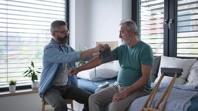 Son measures fathers blood pressure using a home blood pressure monitor. Senior man recovering after surgery, son helping him to return to daily activities.
