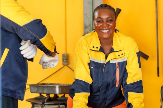 African female mechanic smiling in garage. Wearing blue and yellow uniform, showing positivity and job satisfaction. tools and equipment, professionalism, and successful automotive repair work.