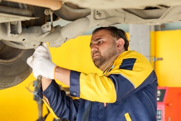 Mechanic working under car, wearing white gloves and blue-yellow uniform. Focused on detailed...