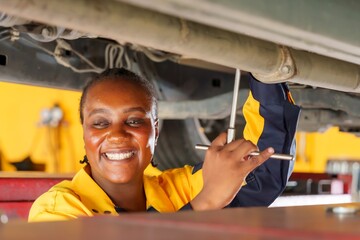 African female mechanic smiling while working under car. Wearing blue-yellow uniform. Represents...