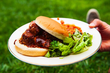 A hand holds a white paper plate with salad, baked beans, and an overflowing barbecue pork sandwich at an outdoor picnic