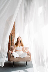 A young female yogi in white clothes sits in the lotus position on a chair against a background of...
