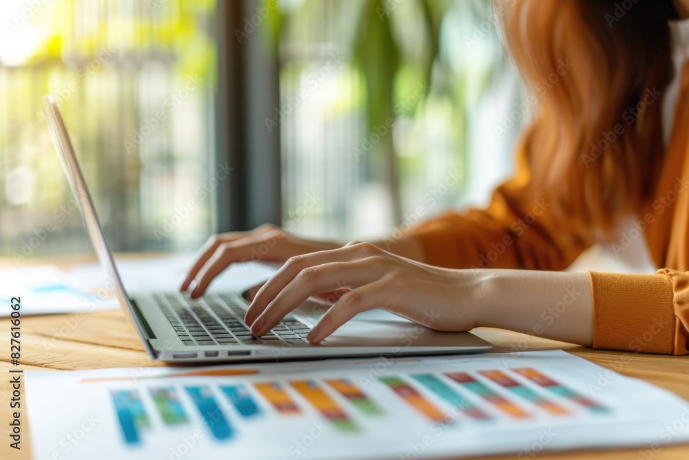 Poster a woman is typing on a laptop in front of a table with several papers. the papers have graphs on the