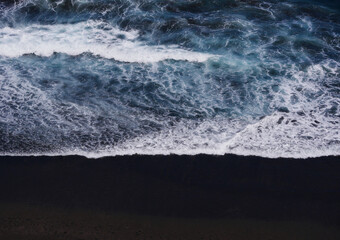Aerial photo of a sea water surface. Blue water with foam on waves. Beach with black volcanic sand - view from above. Stylized background abstract texture photo. Tenerife, Puerto de la Cruz
