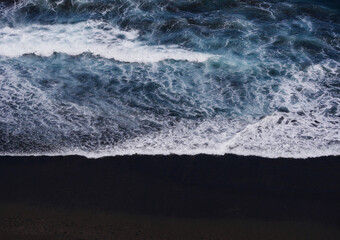 Aerial photo of a sea water surface. Blue water with foam on waves. Beach with black volcanic sand...