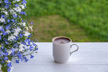 Mug of coffee on a white table in balcony near by lobelia in a pot on blurred background of green...
