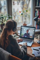 Woman with headphones having a video conference on laptop while working from home at a desk near the window with natural light and plants.