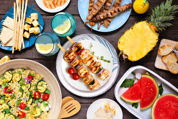 Summer food table scene with  meat skewers, salad, fruit and snacks. Above view on a dark wood background.
