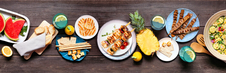 Summer food table scene with  meat skewers, salad, fruit and snacks. Top down view on a dark wood...