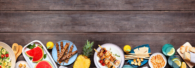Summer food bottom border with  meat skewers, salad, fruit and snacks. Top view on a dark wood...