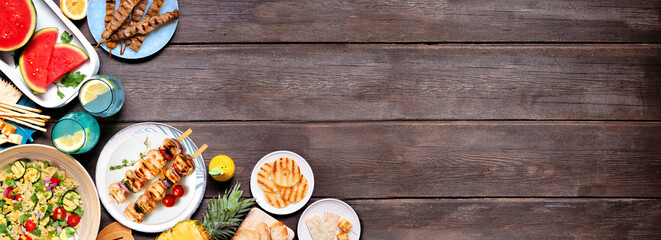 Summer food corner border with  meat skewers, salad, fruit and snacks. Overhead view on a dark wood banner background. Copy space.