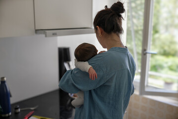 Mother in blue shirt holding baby in kitchen, balancing parenting and household chores, showcasing...