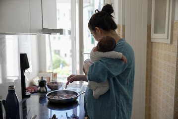 Mother holding baby while cooking in kitchen, demonstrating multitasking skills by managing meal prep and childcare, highlighting the daily balance of motherhood and household responsibilities