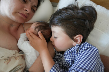 Mother and two young children lying together on a bed, baby nursing and older sibling tenderly holding baby’s head, family bonding moment, relaxed atmosphere, intimate and serene scene