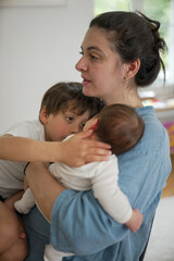 Mother holding baby while young child eats at the table, representing the multitasking nature of parenting and the daily balance of family life, filled with love and care for both children