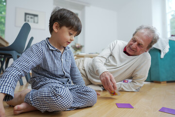Fototapeta premium Elderly man in beige and young boy in checkered pajamas playing cards on the living room floor, enjoying quality time together, focus on their expressions and the joy of the game