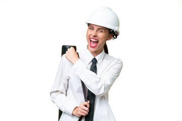 Young architect woman with helmet and holding blueprints over isolated background celebrating a victory