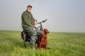 An elderly cyclist pauses in the early morning on a green meadow. He sits on the luggage rack, his...