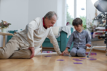 Elderly man and young boy playing card game on living room floor, boy enthusiastically placing...