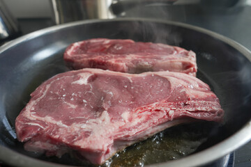 Two raw ribeye steaks searing in a hot pan, emitting steam, capturing the initial stages of...