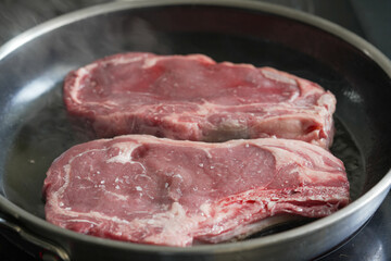 Two raw ribeye steaks searing in a hot pan, emitting steam, capturing the initial stages of cooking, suitable for culinary food preparation, and steak recipe content