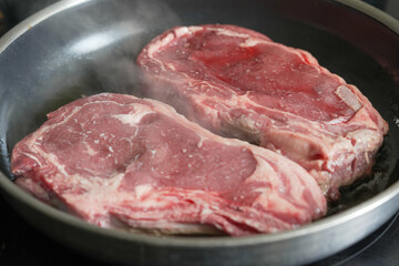 Two raw ribeye steaks searing in a hot pan, emitting steam, capturing the initial stages of cooking, suitable for culinary food preparation, and steak recipe content