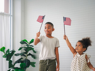 Two young kids with the flag of United States standing in the living room at home while celebrating...