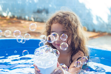 Uma criança dentro de uma piscina infantil azul, brincando, com bolhas de sabão ao redor, no...