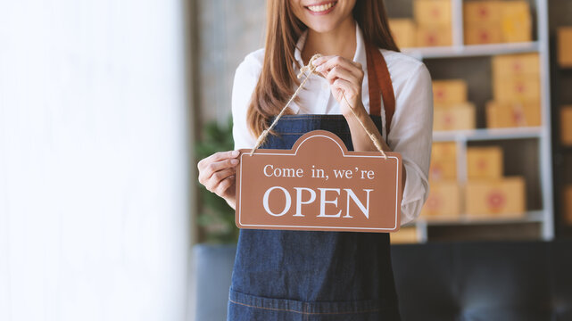 A female shop owner holding an Open sign with a smile, inviting customers to enter her store.