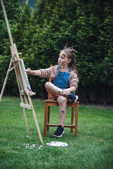 European brunette girl sitting on chair painting on easel on green grass background in nature artist