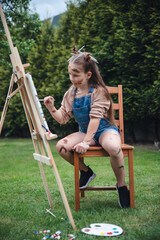 Street artist with easel girl 8 years old European appearance jumpsuit with paints drawing a picture