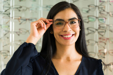 Cheerful young woman tries on new eyeglasses with a wall of frames in the background at her local...