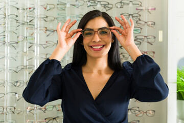 Smiling woman trying on new eyeglasses with many frames on display in the background at an ophthalmologist's or optician's office