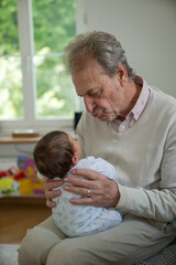 Obraz premium Grandfather lovingly cradling his newborn grandchild, both in a light, airy room. The baby wears a light blue onesie, while the grandfather is dressed in a light sweater