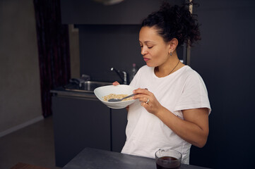 Happy African American woman in pajamas, holding a muesli bowl in the home kitchen. Healthy...
