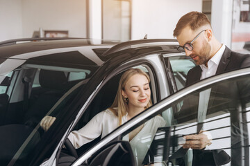 Happy woman with car dealer in auto show or salon. Car Sales Manager Showing Auto To Lady Buyer Sitting In Luxury Automobile Dealership Store.