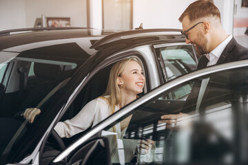 Happy woman with car dealer in auto show or salon. Car Sales Manager Showing Auto To Lady Buyer Sitting In Luxury Automobile Dealership Store.